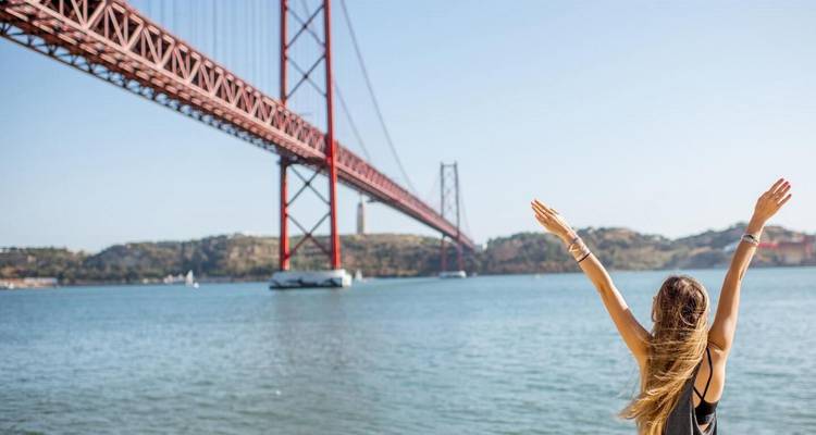 Joyful visitor raises arms in front of Lisbon's 25 de Abril suspension bridge on a clear day.