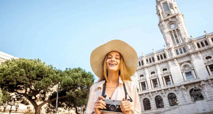 Young tourist holding a camera smiles in front of Porto City Hall tower under blue skies.
