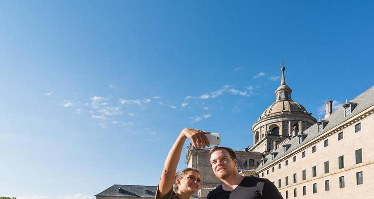 Un jeune couple en train de prendre un selfie avec le monastère royal aux dômes de l'Escorial en arrière-plan et un ciel bleu dégagé