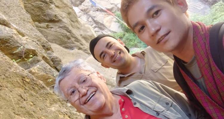 Selfie van drie glimlachende reizigers tegen een rotsachtige klifachtergrond met een gebedsvaandel erboven.