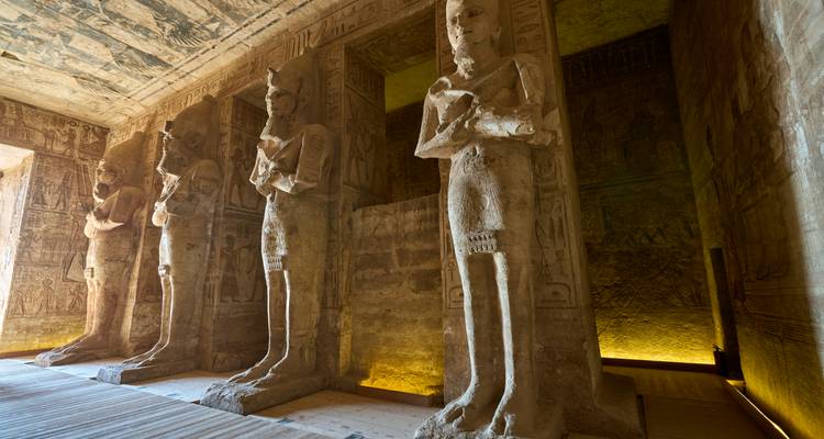 Rangée de statues pharaoniques colossales à l'intérieur du sanctuaire du temple faiblement éclairé avec des murs sculptés.