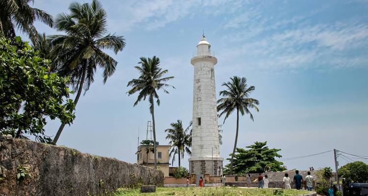 Faro blanco rodeado de palmeras con visitantes caminando alrededor de su base en el Fuerte de Galle.