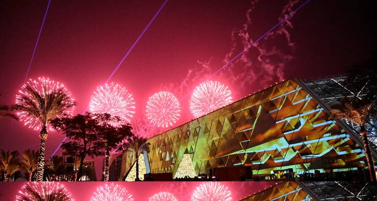 Façade du Grand Musée égyptien illuminée par des projections colorées avec des feux d'artifice spectaculaires au-dessus reflétés sur une surface mouillée