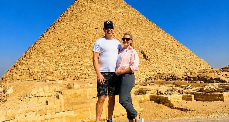 Ein lächelndes Paar posiert vor einer riesigen Pyramide unter einem klaren blauen Himmel in der Wüste.