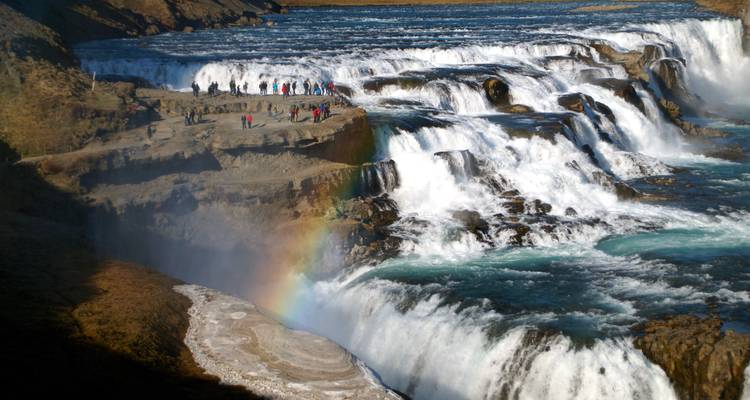 Krachtige meertrapige Gullfoss waterval met regenboog, toeristen die op een rotsachtige uitkijkpunt staan.