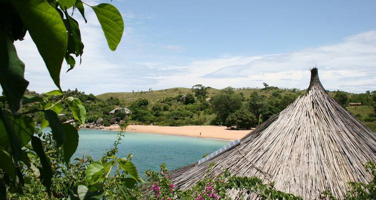 Cabane au toit de chaume surplombant un lac turquoise et une plage de sable fin encadrés par une verdure luxuriante.