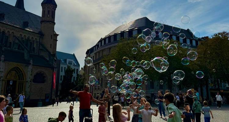 Un artista callejero crea enormes burbujas de jabón que deleitan a un gentío de niños y adultos en una plaza europea soleada.