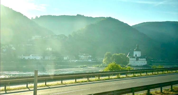 Carretera panorámica que corre junto a un río con un castillo en una isla respaldado por colinas verdes y brumosas bajo una brillante luz matutina.