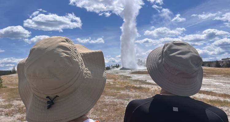 Les voyageurs coiffés de chapeaux de soleil observent l'geyser Old Faithful entrer en éruption sur un fond de ciel bleu