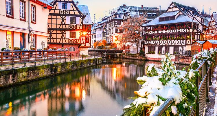 Las casas de entramado de madera cubiertas de nieve se reflejan en el tranquilo canal de la Petite France de Estrasburgo al atardecer.
