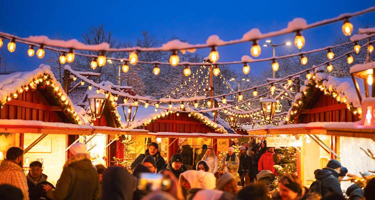 Mercado navideño cubierto de nieve y lleno de gente, iluminado por hileras de luces cálidas y mágicas al atardecer.
