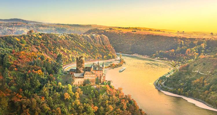 Vista aérea dorada de un castillo medieval encaramado sobre el sinuoso Desfiladero del Rin rodeado de follaje otoñal.
