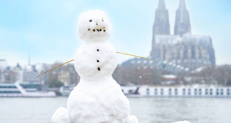 Un muñeco de nieve adorable está de pie a la orilla del río con las borrosas torres gemelas de la Catedral de Colonia de fondo en la nieve.
