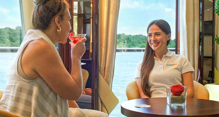 Dos mujeres disfrutan de cócteles en un salón luminoso de crucero fluvial con vistas panorámicas al agua a través de grandes ventanas.