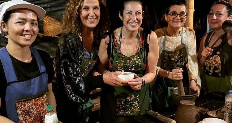 Participantes sonrientes usando delantales estampados en una clase de cocina laosiana por la noche moliendo ingredientes.