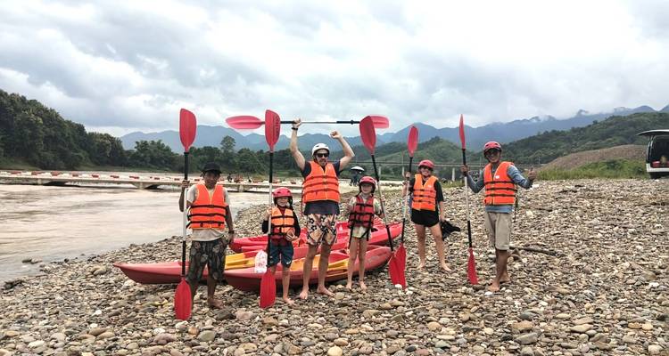 Grupo sonriente en chalecos salvavidas sosteniendo remos junto a kayaks rojos en una playa de guijarros a orillas del río