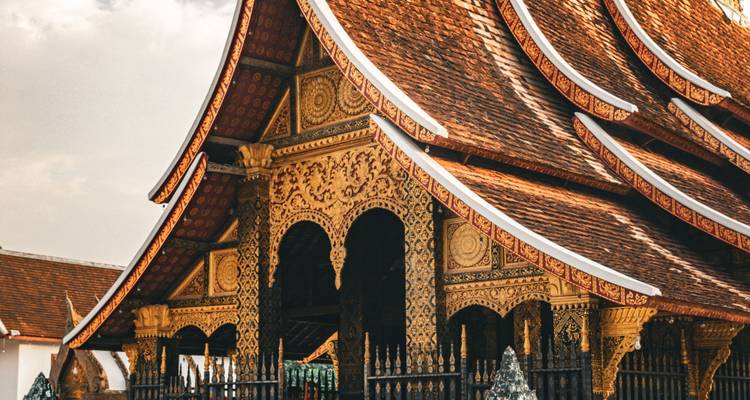 Kunstvoll geschnitzte und vergoldete Dächer und Fassade eines traditionellen laotischen Tempels vor einem sanft beleuchteten Himmel.