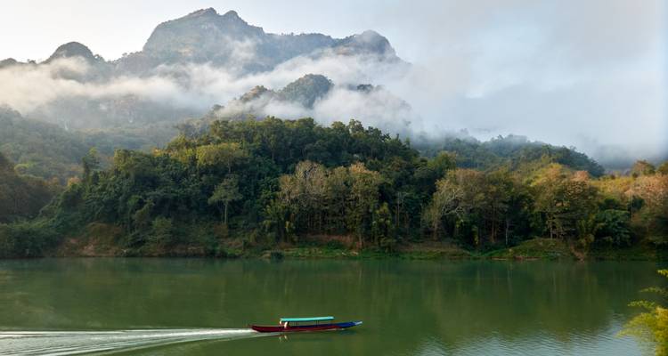 Ein Longtail-Boot gleitet über einen ruhigen grünen Fluss, während nebelverhüllte bewaldete Berge im Hintergrund aufragen.