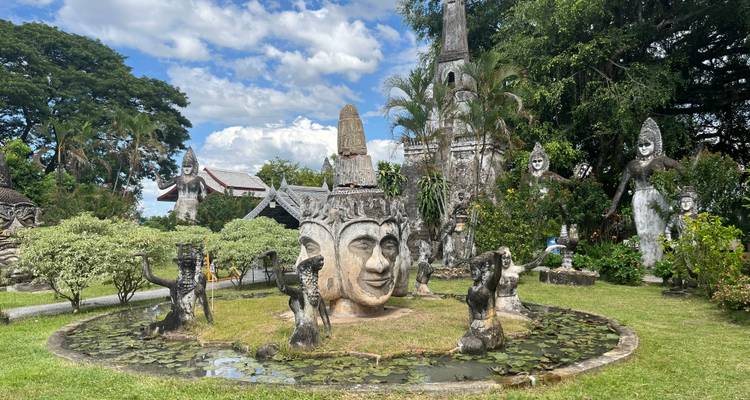 Weitläufiger Blick auf einen Skulpturengarten mit surrealen buddhistischen und hinduistischen Figuren, die einen kleinen Seerosenteich umgeben.