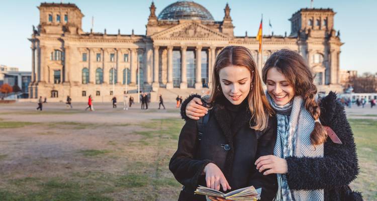 Dos mujeres jóvenes consultan un mapa en el césped frente al edificio del Reichstag de Berlín en una tarde despejada.