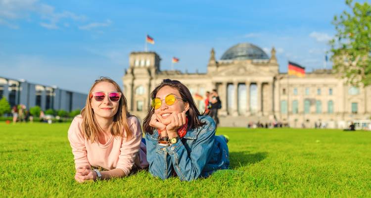 Dos mujeres jóvenes con gafas de sol coloridas relajándose en el césped frente al edificio del Reichstag con cúpula de cristal en Berlín.