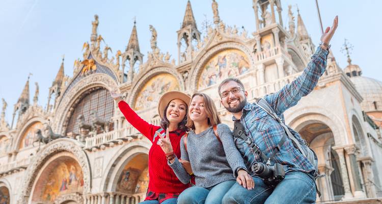 Drei fröhliche Reisende posieren mit erhobenen Armen vor der prunkvollen Basilika San Marco in Venedig.