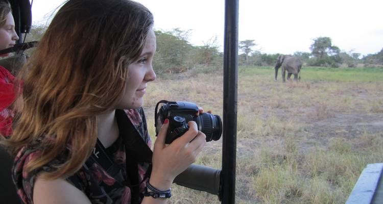 Joven mujer dentro de un vehículo safari abierto enfocando su cámara en un elefante solitario pastando en la sabana.