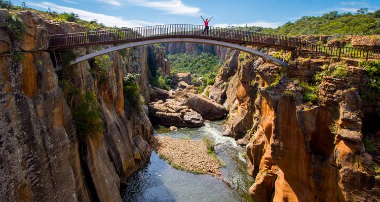 Viajero levantando los brazos en la cima de un elegante puente de arco que cruza un cañón espectacular con paredes de roca en capas y un río caudaloso.