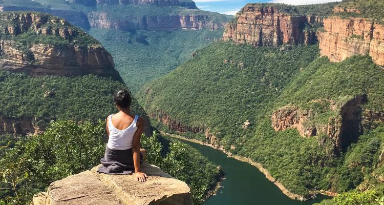 Mujer sentada tranquilamente en el borde de un acantilado mirando hacia el exuberante paisaje verde y el río sinuoso del Cañón del Río Blyde.
