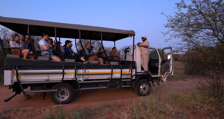 Open-air safari truck where a guide speaks to a seated group of tourists on a dirt road surrounded by bush at dusk.