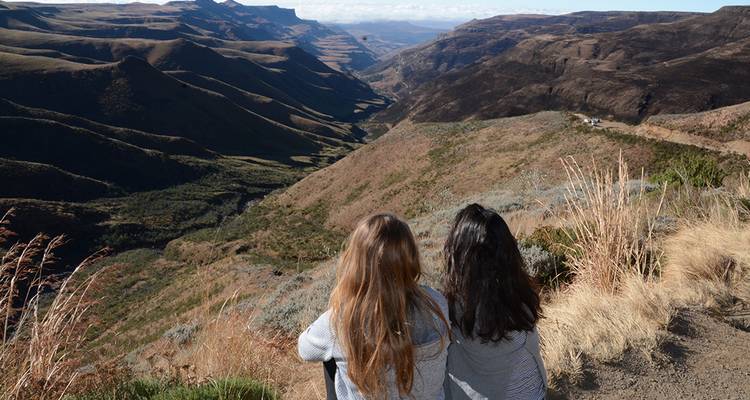 Two young women sit on a mountain lookout taking in a vast rugged valley landscape.