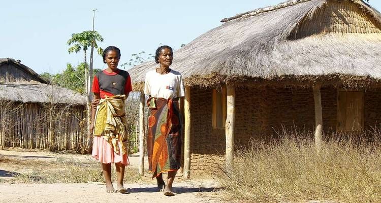 Deux femmes malgaches vêtues de pagnes colorés marchent devant des maisons en terre avec des toits de chaume dans un village rural.