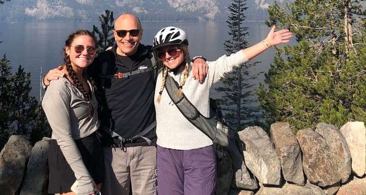 Trois voyageurs heureux portant des casques de cyclisme et des sacs à dos s'embrassent sur un point de vue en pierre surplombant un lac de montagne serein et une forêt de conifères.