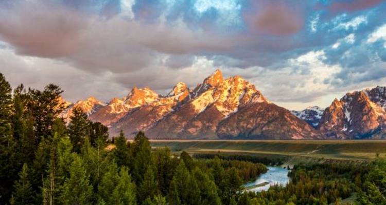 La lumière du coucher de soleil baigne la chaîne de montagnes du Grand Teton aux sommets enneigés au-dessus des forêts de pins et d'une rivière sinueuse.