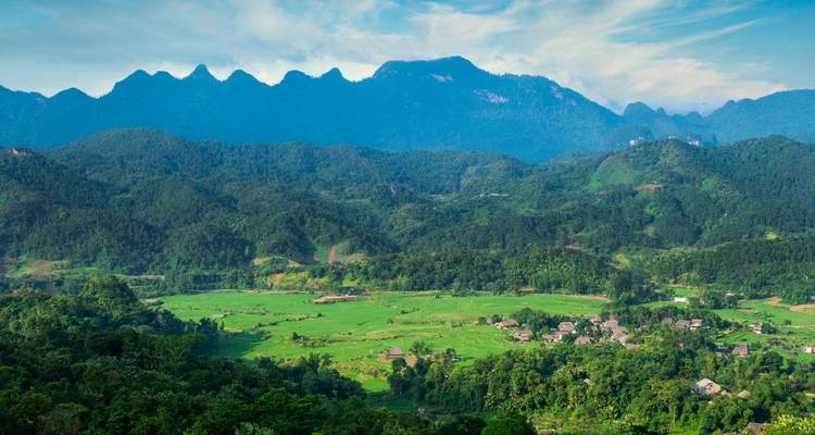 Ein weiter Blick zeigt grüne Täler und ein Dorf unter gezackten blauen Bergen unter einem hellen Himmel.