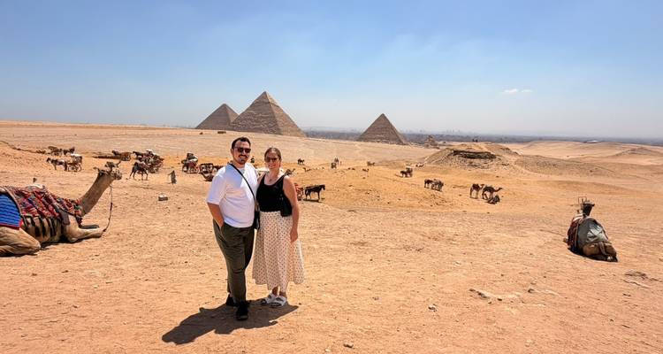 Un couple se tient dans le désert avec les pyramides emblématiques de Giza et des chameaux au repos dispersés dans le paysage sablonneux.