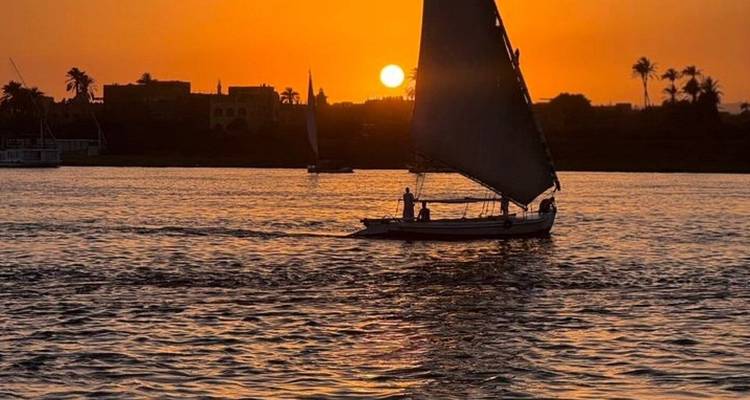Een felucca met een donker zeil glijdt bij zonsondergang over de Nijl, afgetekend tegen een vuuroranje lucht met de zon laag aan de horizon.