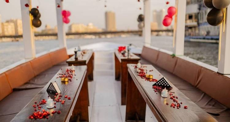 Lange houten banken versierd met rode rozenblaadjes en kaarsen staan opgesteld op het openluchtdek van een boot die langs een stadsrivier vaart.