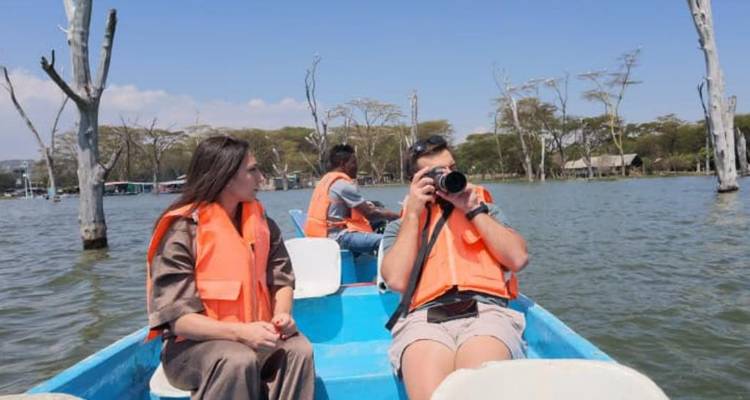 Tres viajeros vistiendo chalecos salvavidas naranjas viajan en un pequeño bote azul en un lago tranquilo salpicado de troncos de árbol desnudos, uno de ellos tomando fotos.