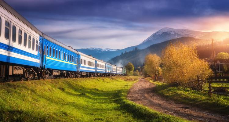 Blue passenger train curving through a lush green valley with snow-capped mountains in warm golden light