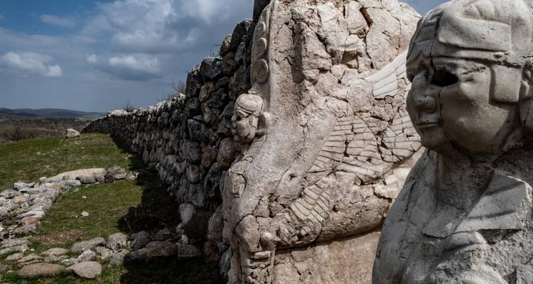 Ancient stone wall with weathered Hittite reliefs and statues under a moody sky on a grassy plain