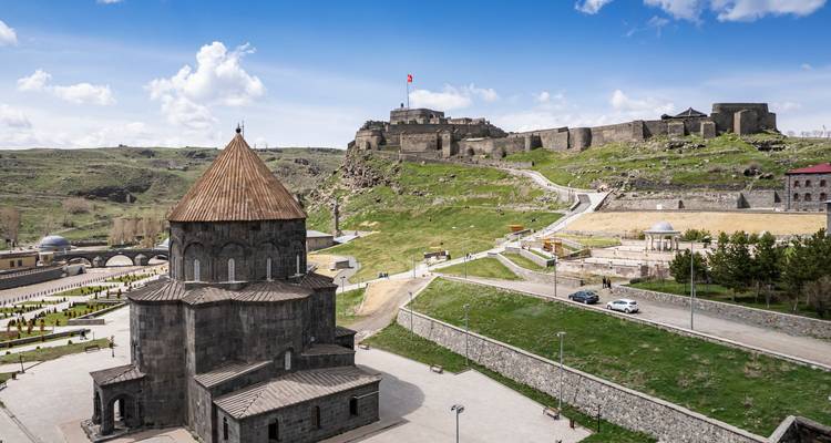 Elevated view of a stone cathedral and Kars Castle set on rolling green hills under a bright blue sky