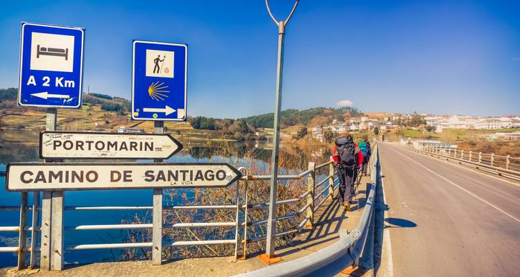 Backpackend pelgrims steken een brug over terwijl ze langs borden van Camino de Santiago en Portomarin lopen naast een rivier.