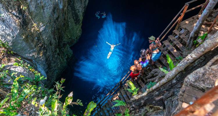 Les voyageurs observent un plongeur qui se jette dans un cenote bleu luminescent depuis des escaliers en bois.