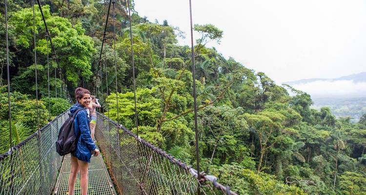 Two hikers smile while crossing a high canopy bridge above misty rainforest valleys.