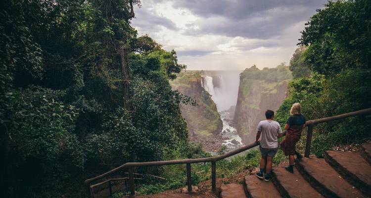Couple stands on a forested path overlooking the dramatic gorge and spray of Victoria Falls.