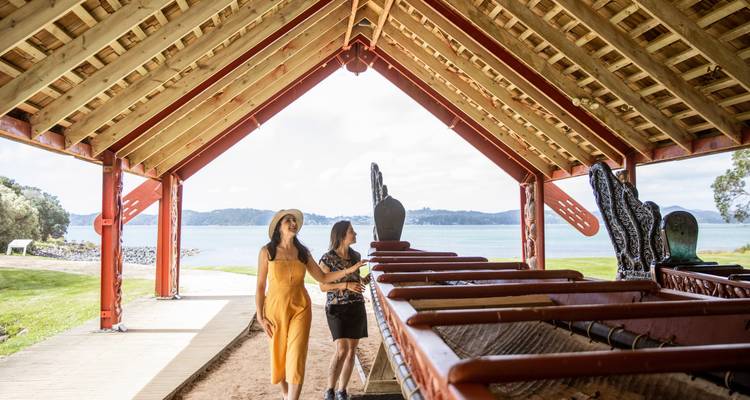 Dos mujeres caminan pasando un refugio de canoa de guerra maori tallada que da vista a una bahía tranquila