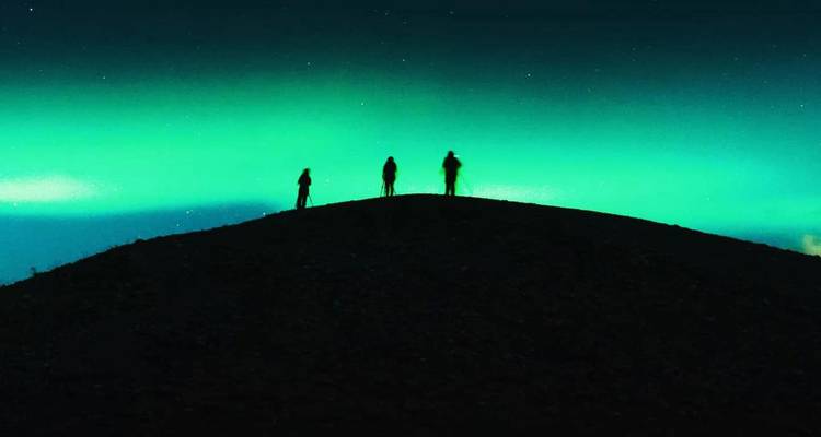 Silhouetten van drie mensen die op een heuvel staan onder het levendige groene Noorderlicht