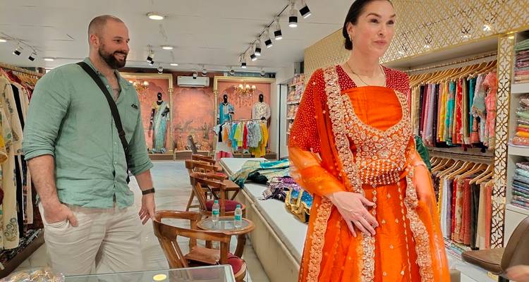 Een vrouw past een felloranje traditionele jurk in een kleurrijke Indiase kledingwinkel terwijl een man in de buurt toekijkt.