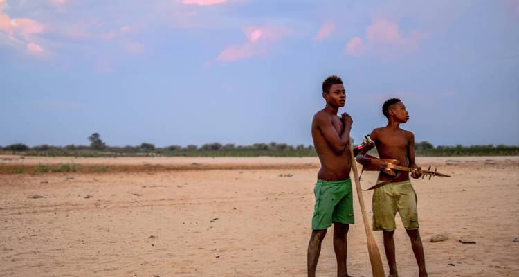 Dos chicos locales parados en una inmensa llanura arenosa al atardecer, uno sosteniendo un instrumento de cuerda hecho a mano.
